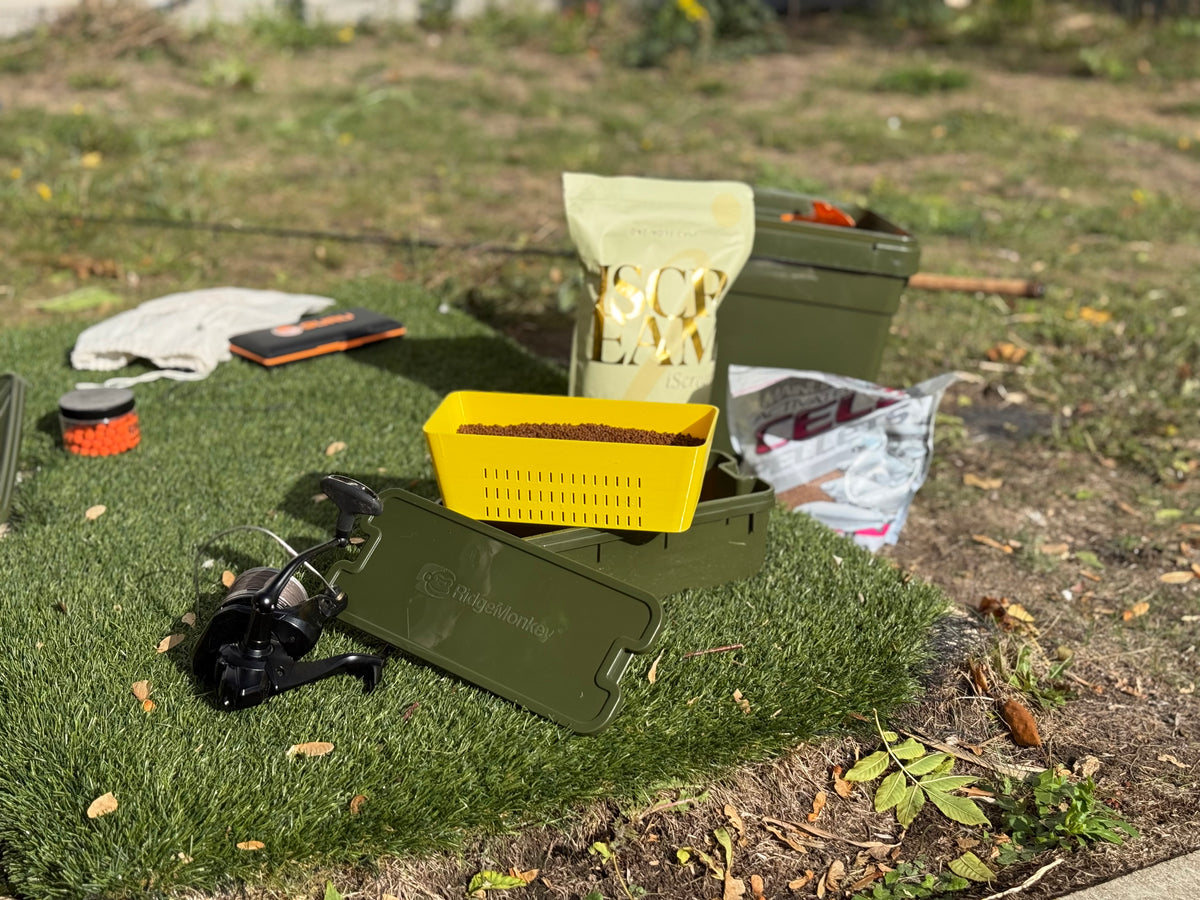 Outdoor fishing tackle setup on grass, featuring an olive green hard case with a yellow tray filled with bait, a black spinning reel, a bag of Sog Earm bait, and other fishing accessories.